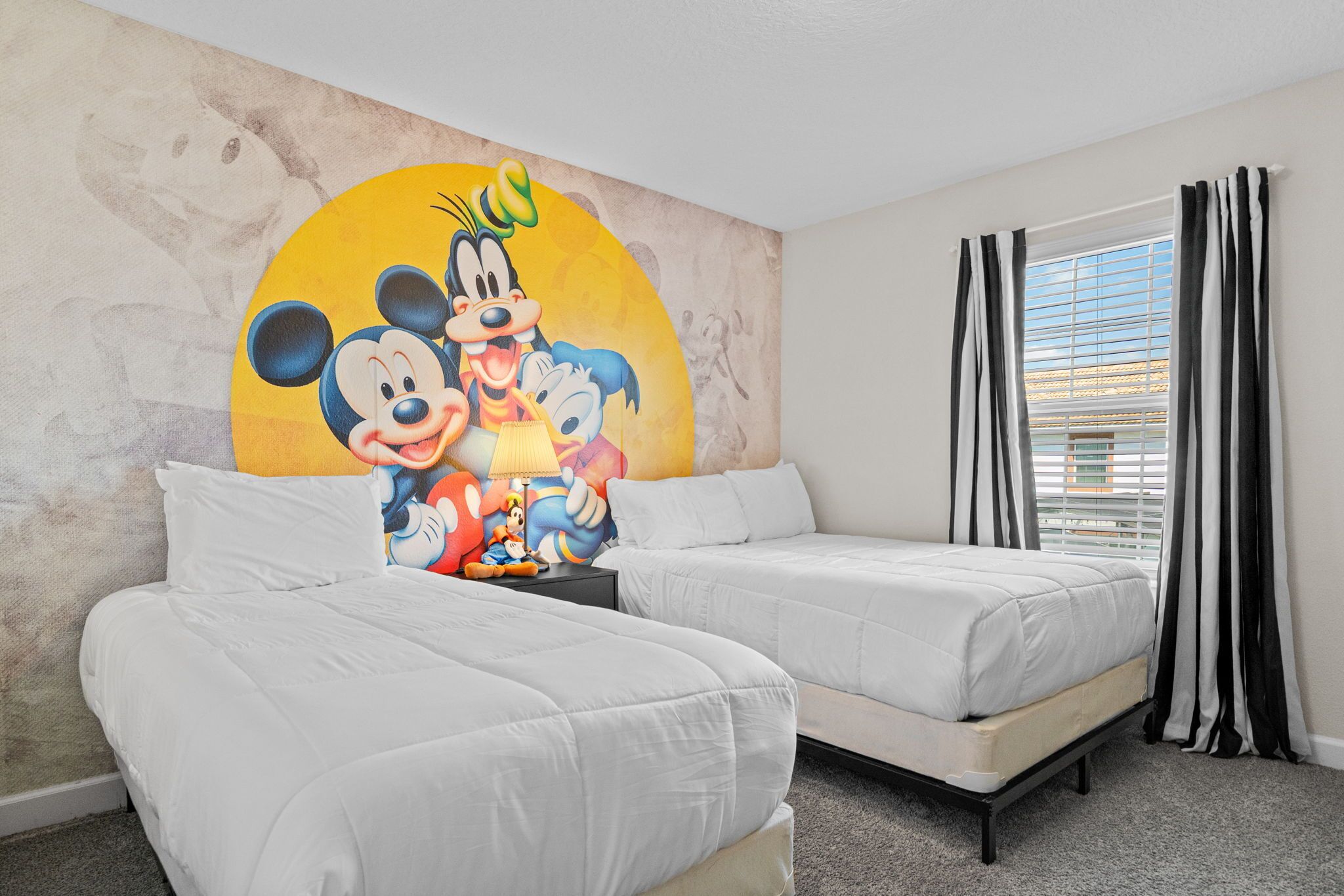 A hotel room with two beds, a Mickey Mouse mural on the wall, and a window with curtains.