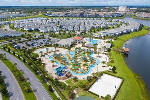 Vista aerea di una zona residenziale con un ampio complesso di piscine, circondato da case unifamiliari e un corpo d'acqua.