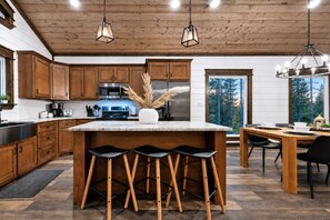 Eine moderne Küche mit Holzausstattung, einer zentralen Kochinsel mit Barhocker und einem Essbereich mit Blick auf Bäume.