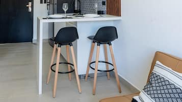 Moderni keittiö, jossa on valkoinen työtaso, kaksi mustaa baarituolia, kattotuuletin ja puinen tehosteseinä.
