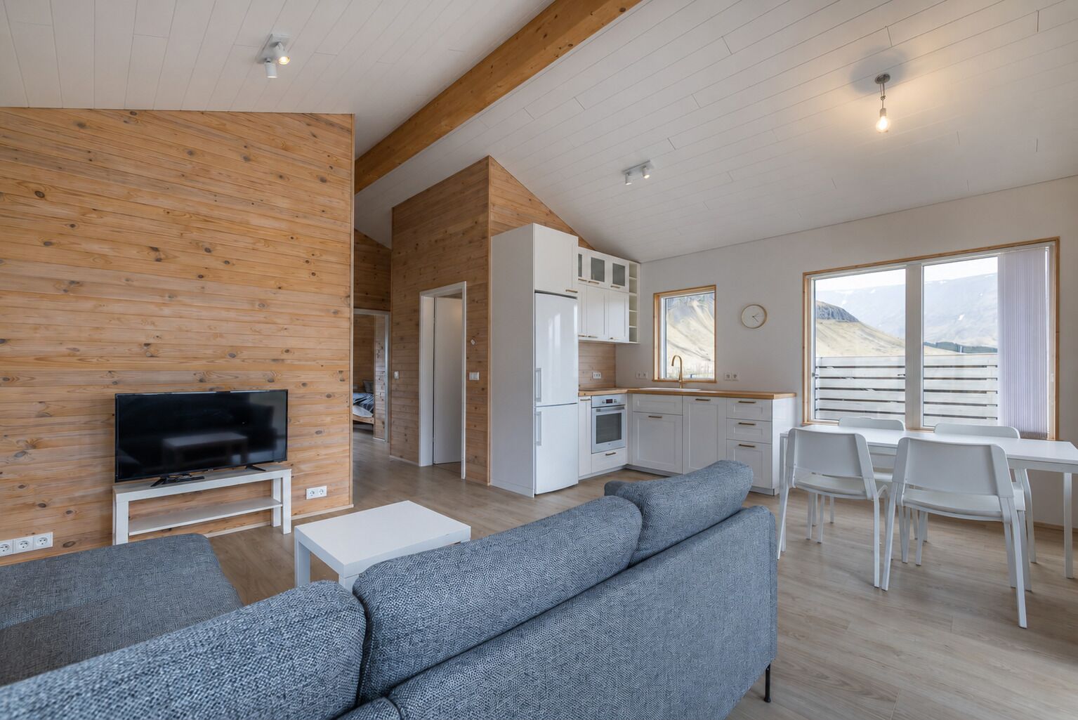 Ein modernes Wohnzimmer mit Flachbildfernseher, grauen Sofas und einem Essbereich mit weißen Stühlen.