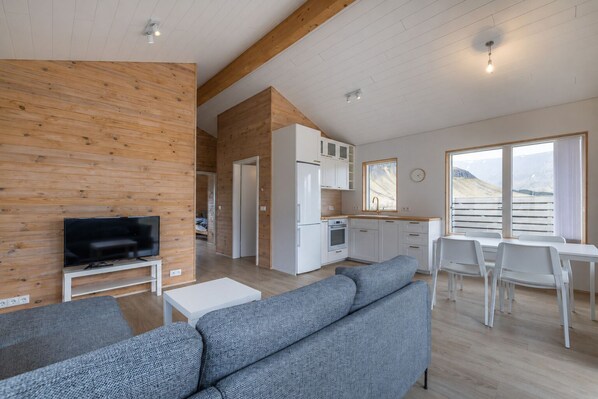 A modern living room with a flat-screen TV, grey sofas, and a dining area with white chairs.