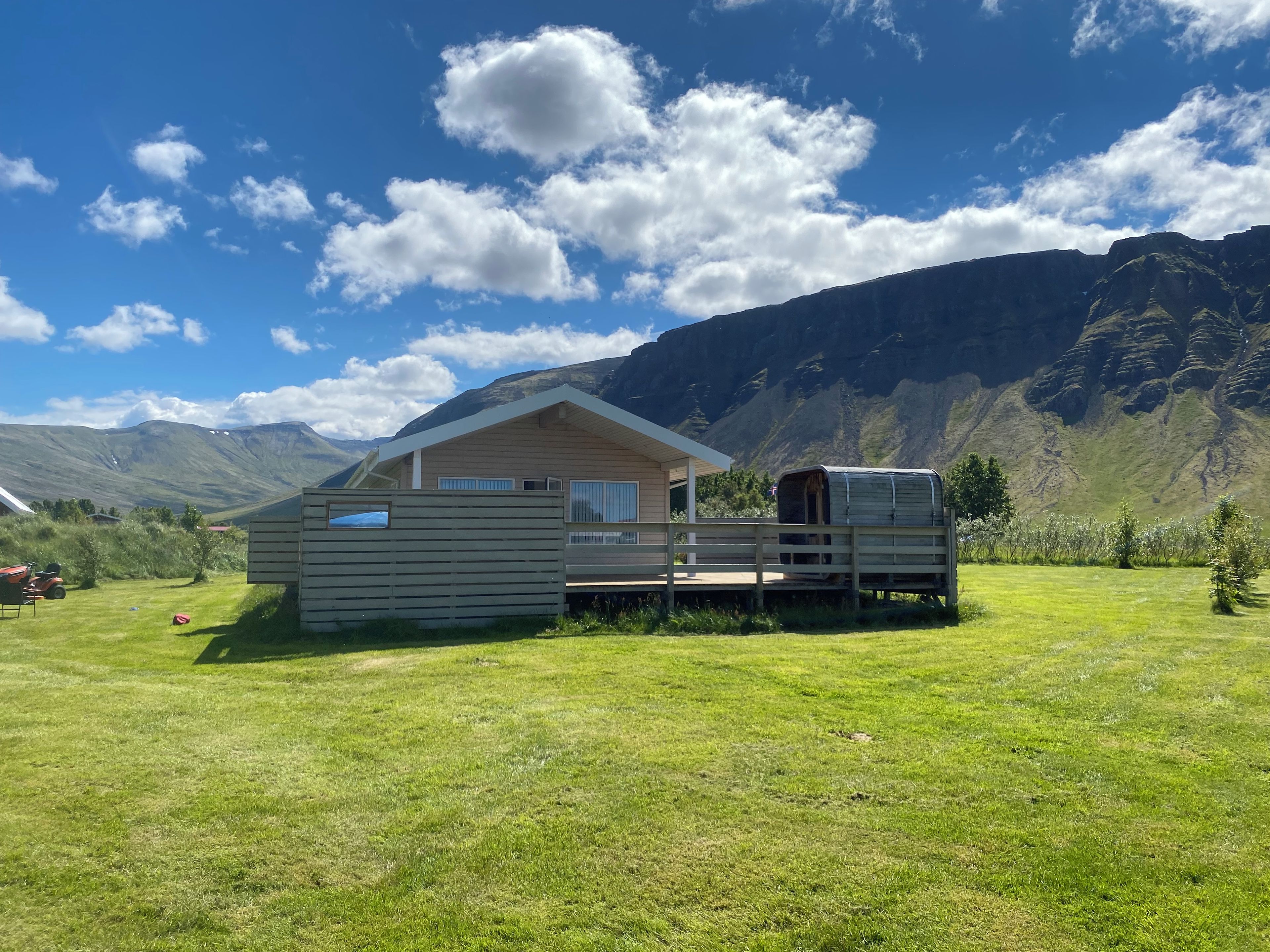 A cabin with a deck and a covered wagon in a grassy field with mountains in the background.