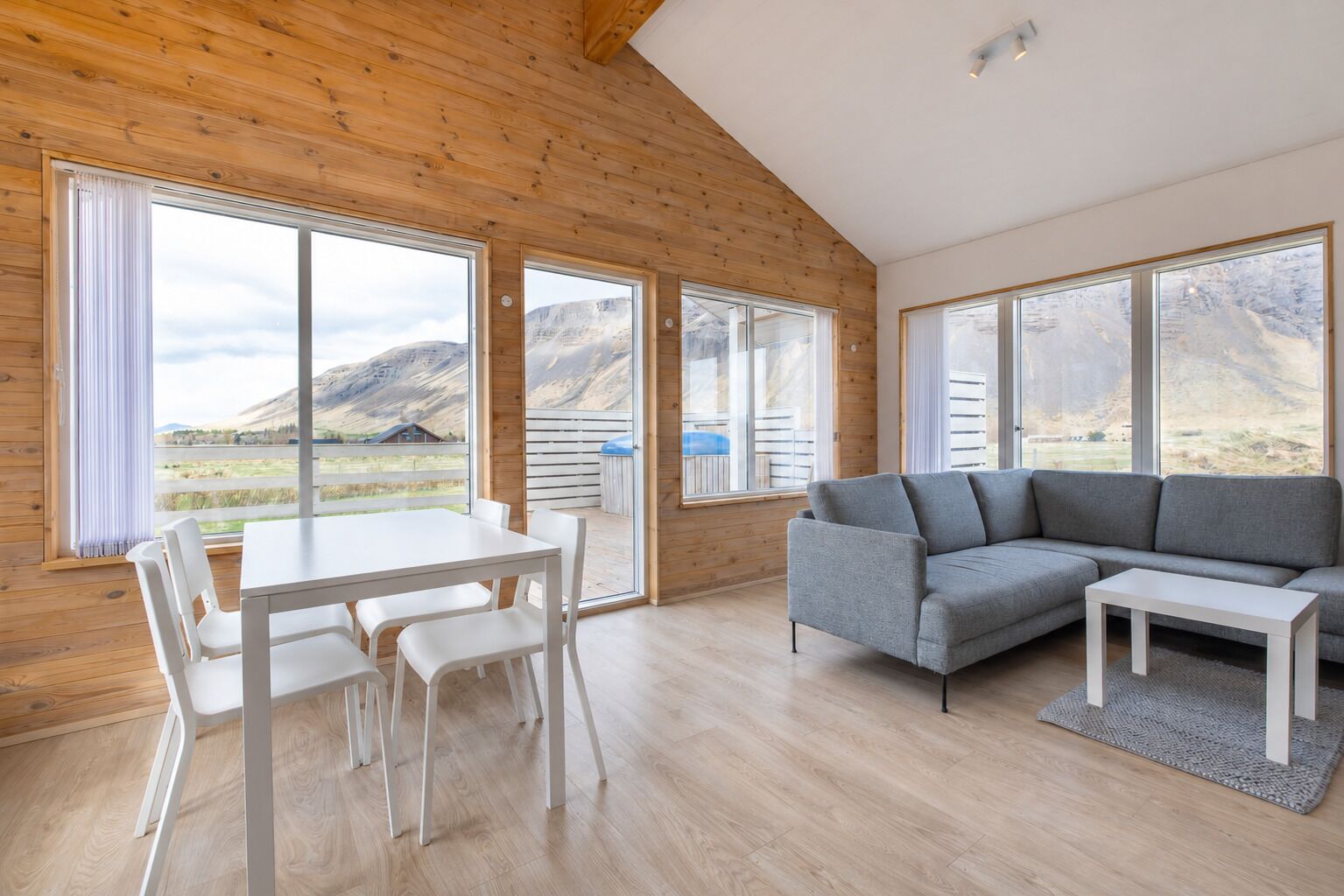 A modern interior with a wooden ceiling, large windows offering a view of a mountainous landscape, a grey sofa, and a white dining table with chairs.