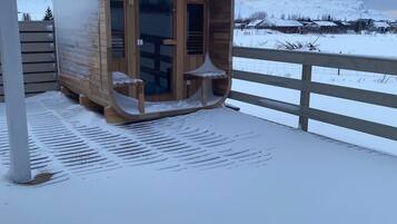 Eine hölzerne Sauna mit Glastüren, auf einer mit Schnee bedeckten Terrasse mit Blick auf eine verschneite Landschaft und Häuser im Hintergrund.