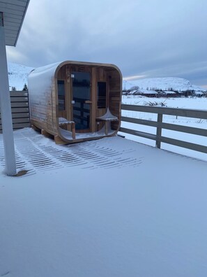 A wooden sauna structure with glass doors, situated on a snow-covered deck with a view of a snowy landscape and houses in the background.