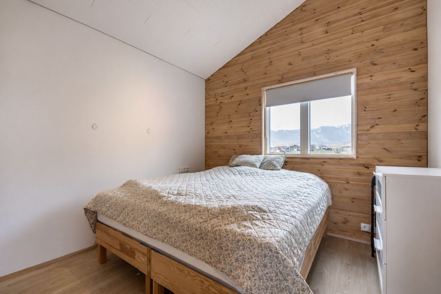 Ein Schlafzimmer mit Holzbalkendecke, einem Bett mit floralem Überwurf, einem Fenster mit Bergblick und einem weißen Gerät.