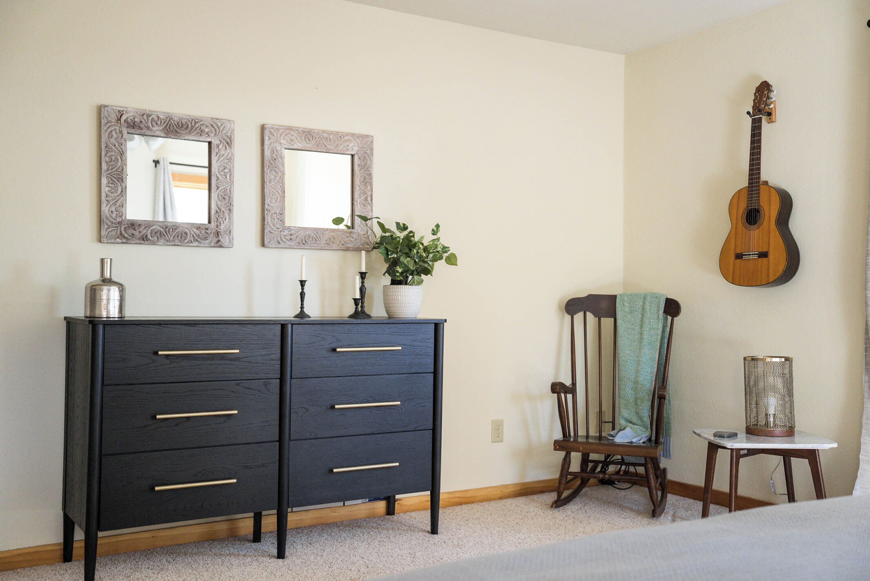 Une chambre avec une commode en bois foncé, deux miroirs muraux, une guitare, une chaise avec une serviette et une petite table avec une lampe.