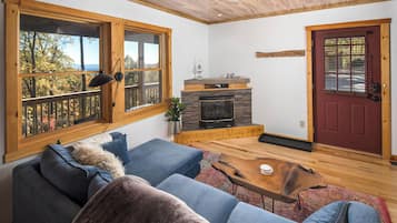 A cozy living room with a fireplace, a sofa, a coffee table, and large windows with a view of trees.