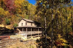A two-story house with a porch and a wooden deck, surrounded by a forest with autumn foliage. - Whisper Ridge 3BR Deep Gap Cabin · Mountain Views · Fire Pit · Sleeps 9 (Watauga County)