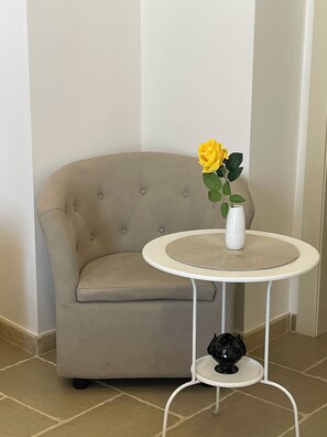 A corner room with a beige sofa, a small round table with a vase and a single yellow flower, and a black decorative object on the table's base. - Aurea Suite - Private Room (Veglie)