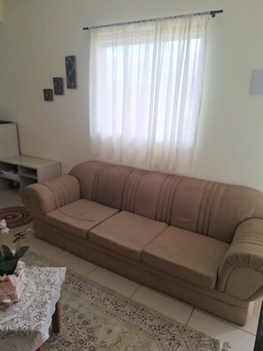 Ein beiges Sofa in einem Wohnzimmer mit einem gemusterten Teppich, einem kleinen Tisch mit einer Spitzenleinentuch und einem Fenster mit transparenten Vorhängen.