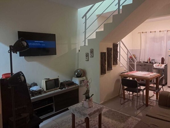 Ein Wohnzimmer mit Fernseher, einem Schreibtisch mit Computer, einer Essecke mit Tisch und Stühlen sowie einer Treppe, die in eine weitere Ebene führt.