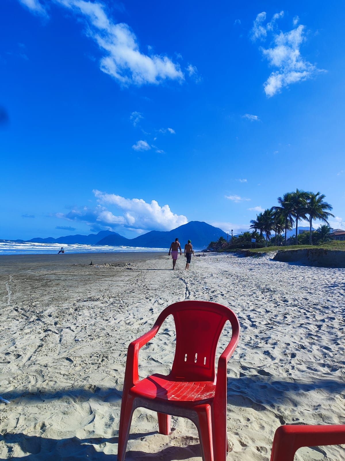 Una playa con una silla de plástico roja al frente, un cielo azul despejado y algunas personas caminando a lo lejos.