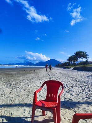 Ein Strand mit einem roten Plastikstuhl im Vordergrund, einem strahlend blauen Himmel und ein paar Personen, die in der Ferne entlanggehen.
