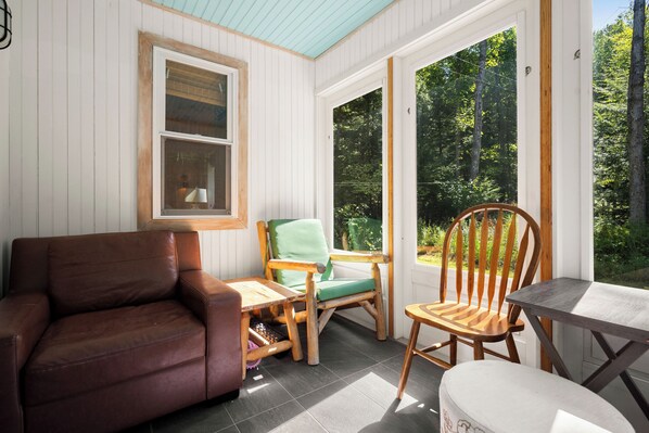 A cozy room with a brown leather sofa, a wooden chair with a green cushion, a small wooden table, and a view of a forest through large windows.