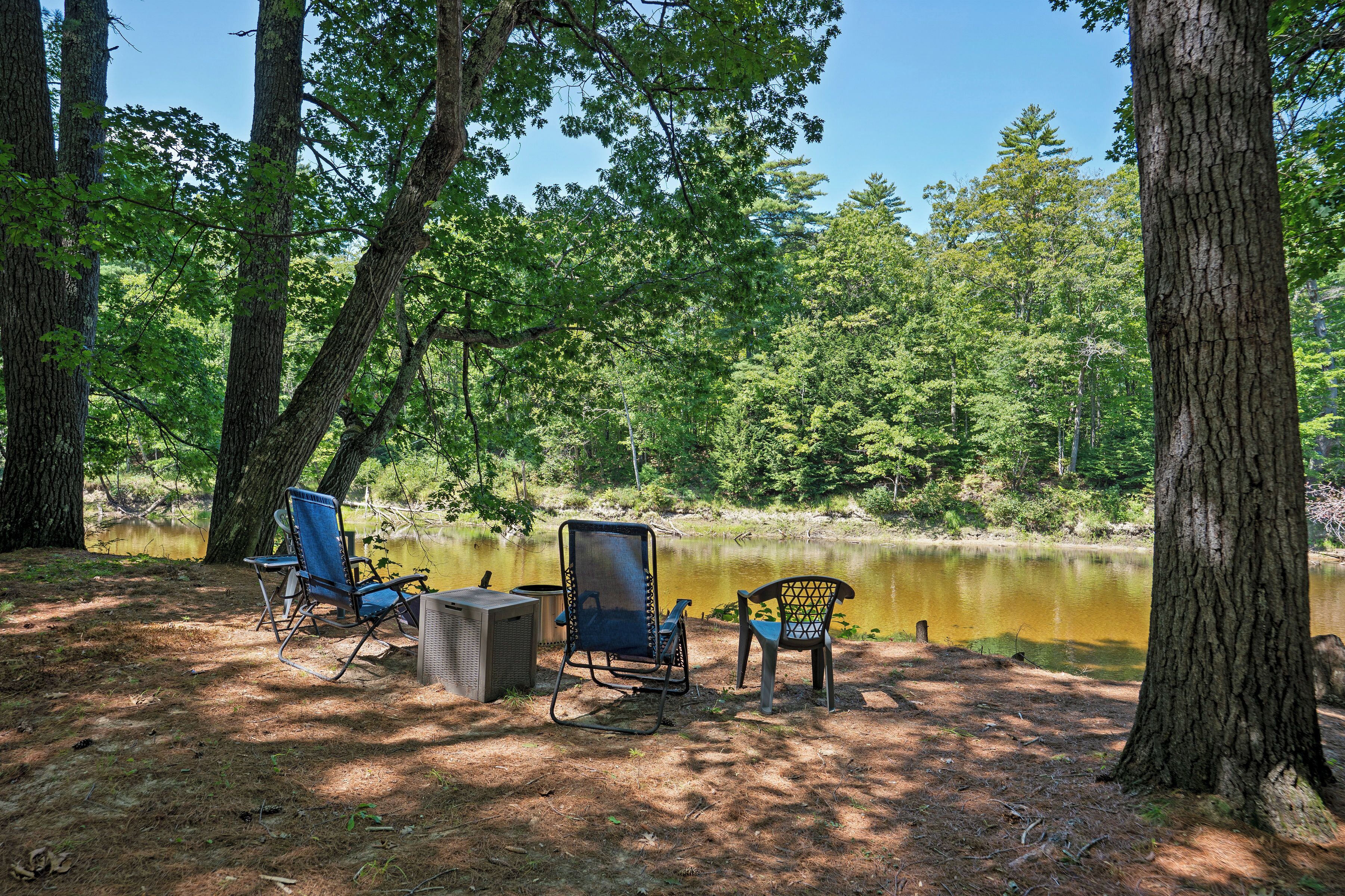 Un terrain de camping avec deux chaises et une table, entouré d’arbres et bordé d’un lac paisible.
