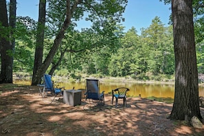 Um camping com duas cadeiras e uma mesa, cercado por árvores e um lago calmo.