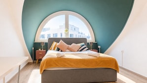 A bedroom with a curved ceiling, a large window, a bed with multiple pillows, a wooden floor, and a fur rug.