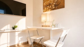 A modern room with a flat-screen TV mounted on the wall, a desk with a lamp, two white chairs, and a small potted plant.