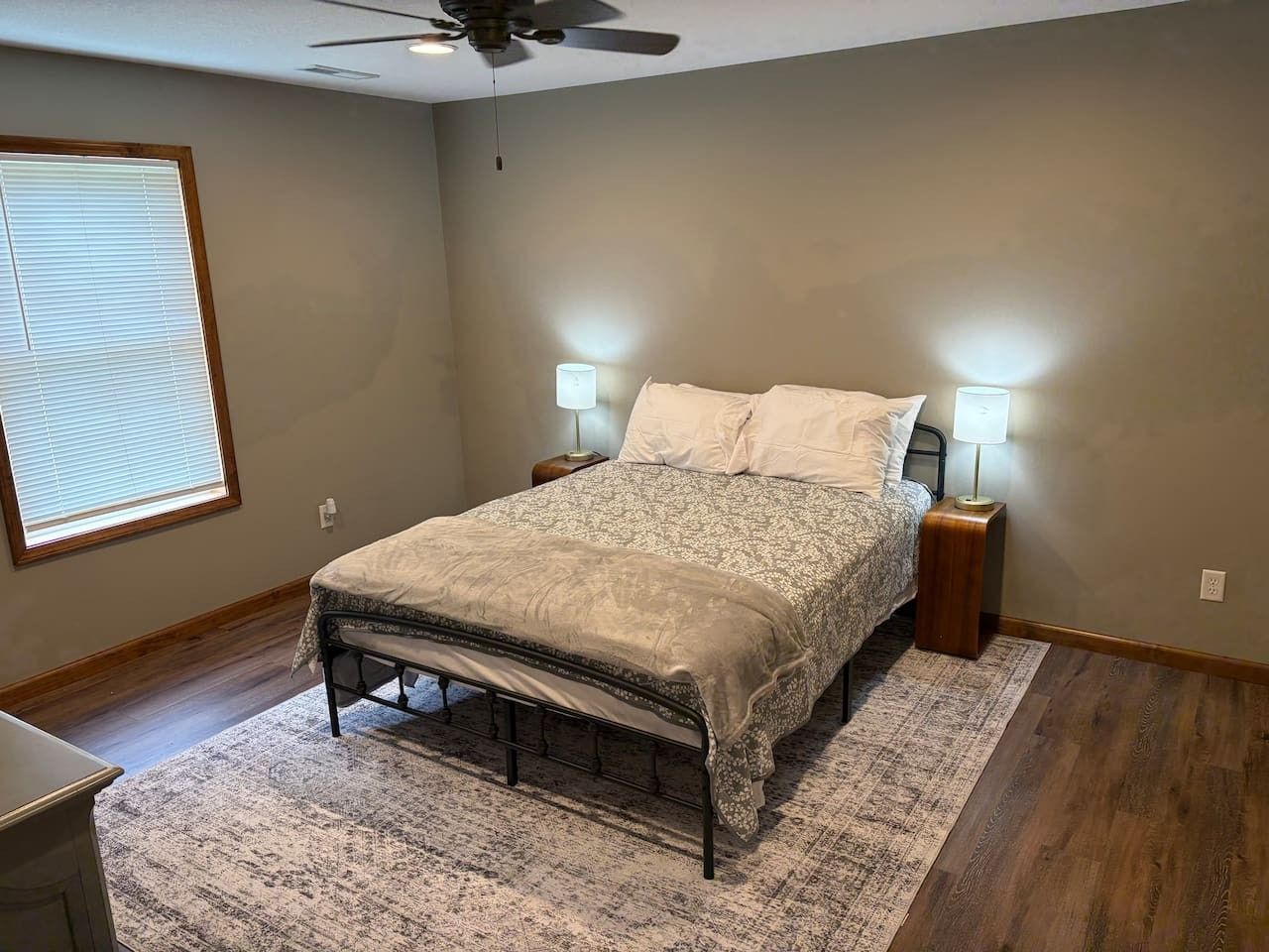 Een slaapkamer met een bed, twee bedlampen, een plafondventilator, een raam met zonwering en een houten vloer.