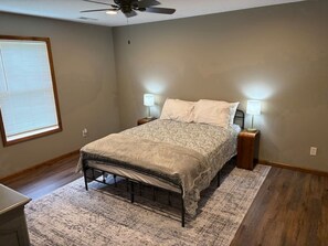 Une chambre à coucher avec un lit, deux lampes de chevet, un ventilateur de plafond, une fenêtre avec des stores et un plancher en bois.