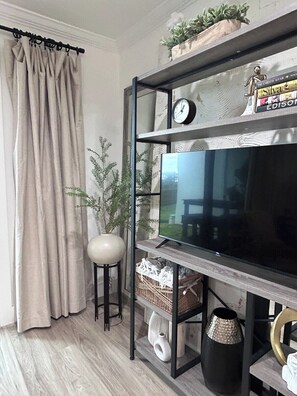 A living room with a TV on a wooden shelf, a potted plant, and a curtain. - New 30A Style! Private Beach | 5 Star Host (Seagrove)