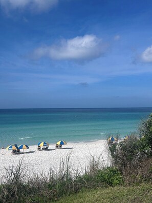Een strand met wit zand en helderblauw water. Er staan meerdere strandparasols en ligbedden.