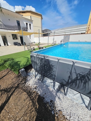 Uma piscina com um deck ao redor, uma área de estar coberta e um edifício moderno ao fundo.