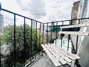 A balcony with a metal railing, a wooden bench, and a view of a cityscape with buildings and trees. - Mons Sanctus - Warrick Ave, a sanctuary in the heavens (London)