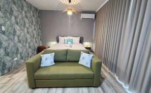 Ein Hotelzimmer mit einem Bett, einem grünen Sofa, einer Wand mit Blättermotiv, einer Deckenleuchte und einer Klimaanlage.