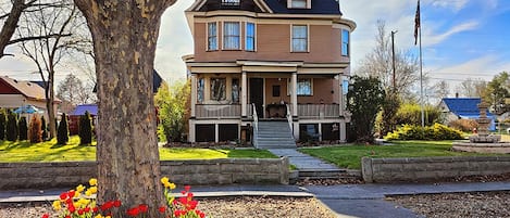 Uma casa de dois andares com varanda frontal, um mastro de bandeira e um jardim com flores vermelhas e amarelas.