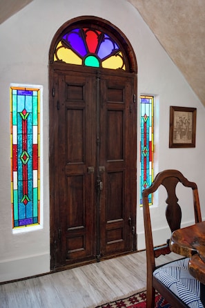 Une pièce dotée d’une porte à double battant en bois, de deux vitraux, d’un tableau encadré accroché au mur et d’une chaise en bois avec un coussin à motifs.