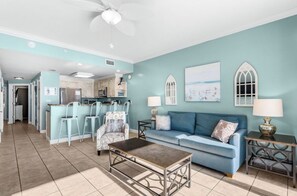 A living room with a blue sofa, a coffee table, and a dining area with chairs and a kitchen in the background. - Panama City Beach Oasis: Panoramic Gulf Views & Direct Beach Access (Panama City Beach)