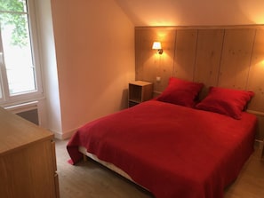 A bedroom with a wooden headboard, a bed with red bedding, a bedside table, and a window with daylight. - The Saddlery (Chaumes-en-Retz)