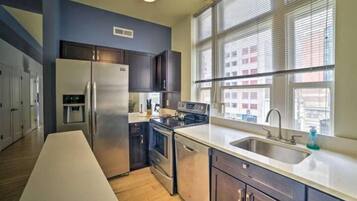 A modern kitchen with stainless steel appliances, a large window with blinds, and a long countertop.