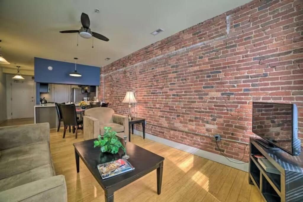 A living room with a brick wall, a ceiling fan, a fireplace, and a television.