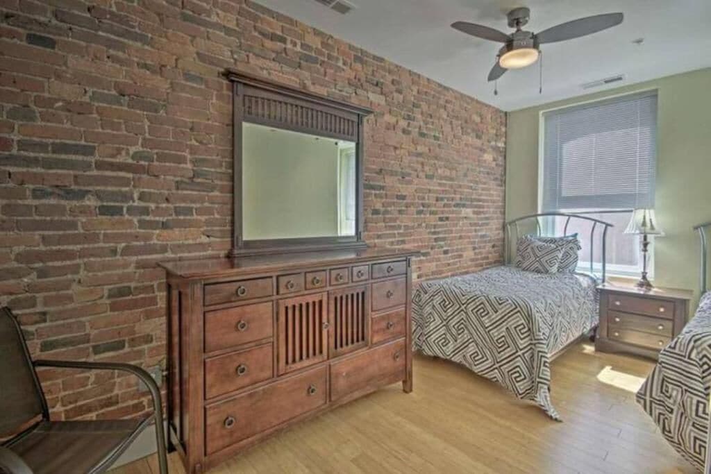 A bedroom with a brick wall, a wooden dresser, a bed with patterned bedding, a nightstand, and a ceiling fan.