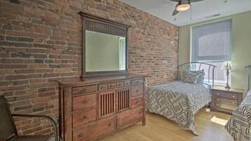 A bedroom with a brick wall, a wooden dresser, a bed with patterned bedding, a nightstand, and a ceiling fan.