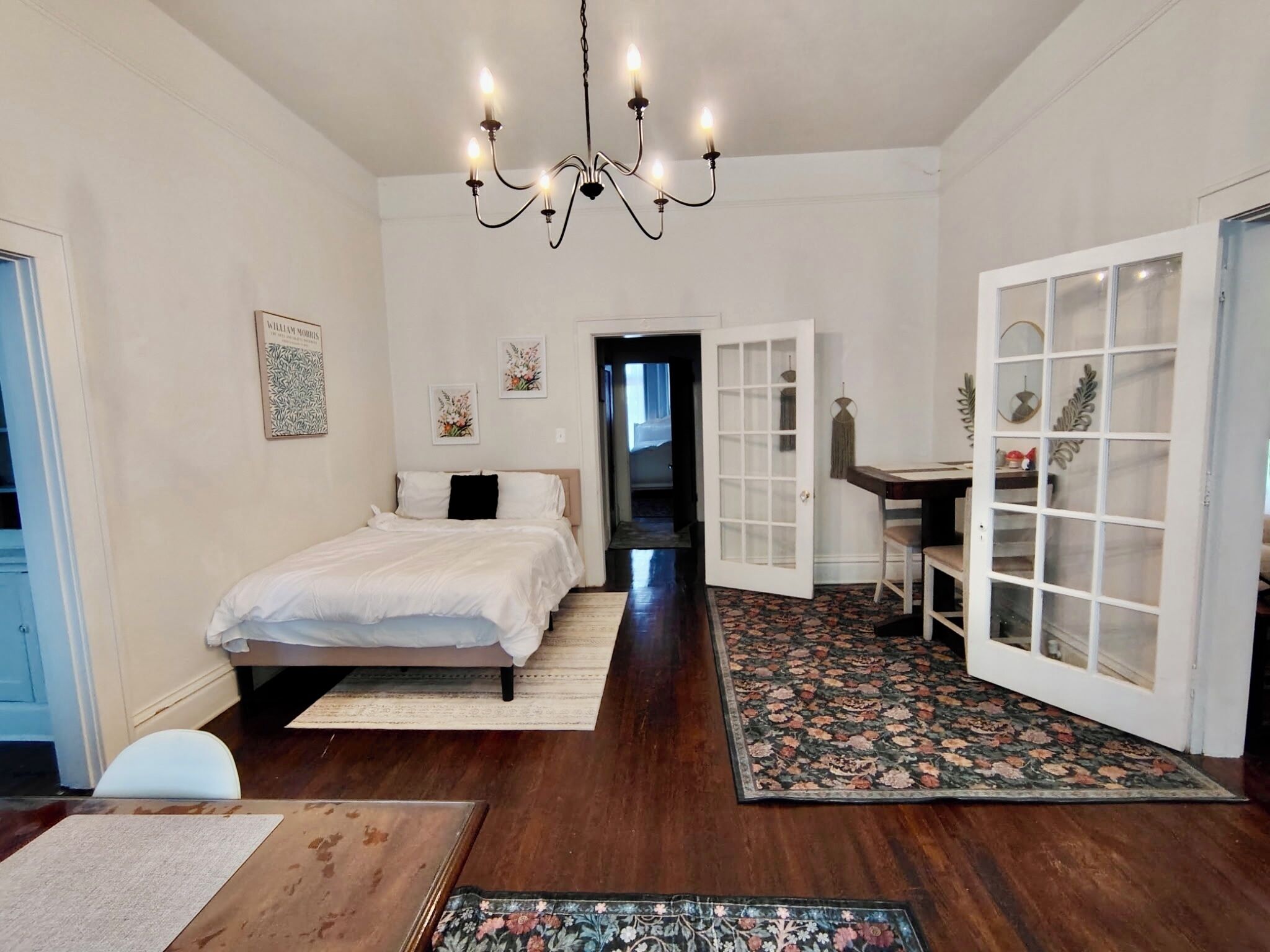 A bedroom with a bed, a desk, and a chair. There is a chandelier, a door, and a window.