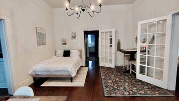 A bedroom with a bed, a desk, and a chair. There is a chandelier, a door, and a window.