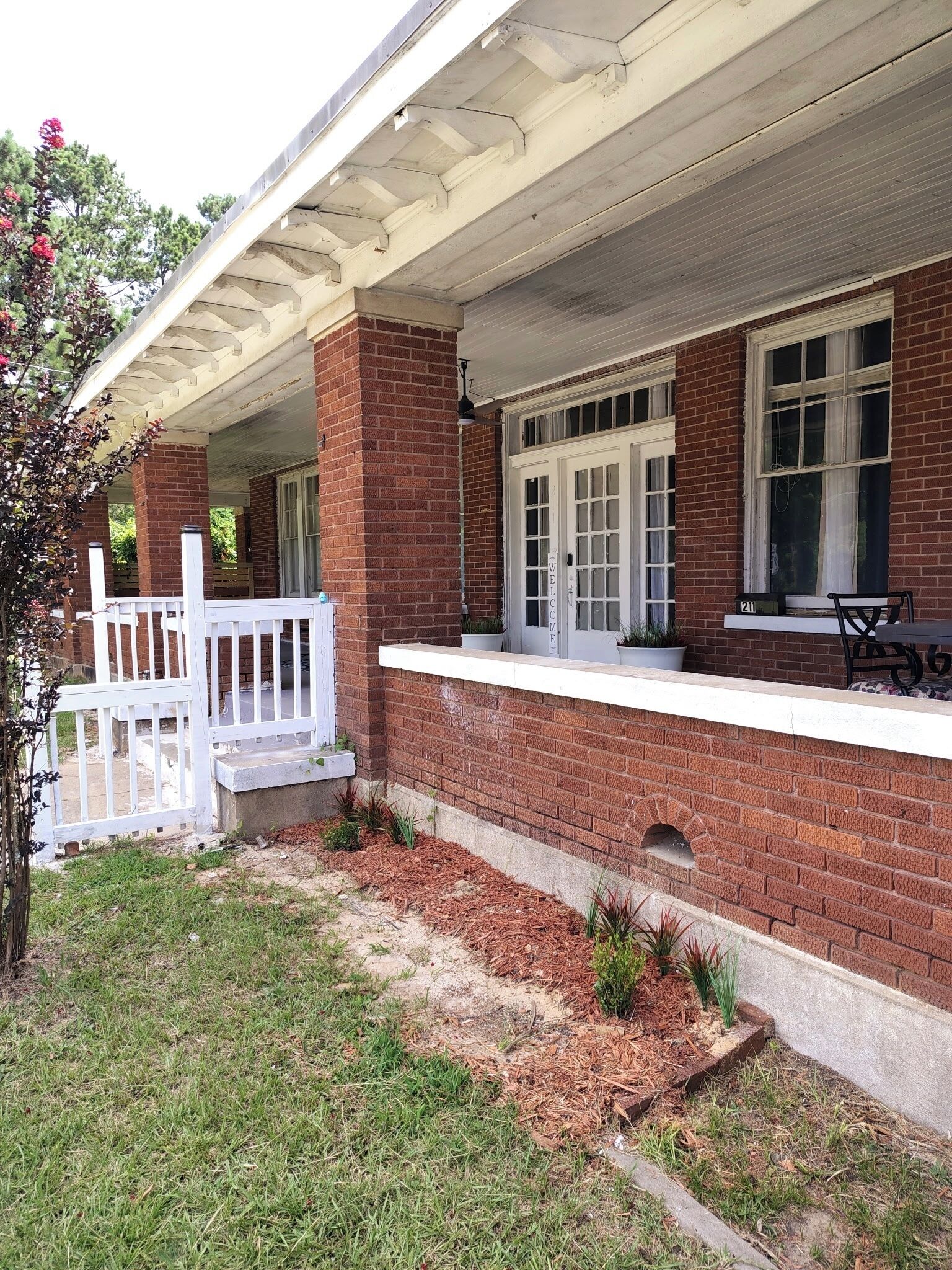 A two-story brick building with a covered porch, a white fence, and a small patio area with a table and chairs.
