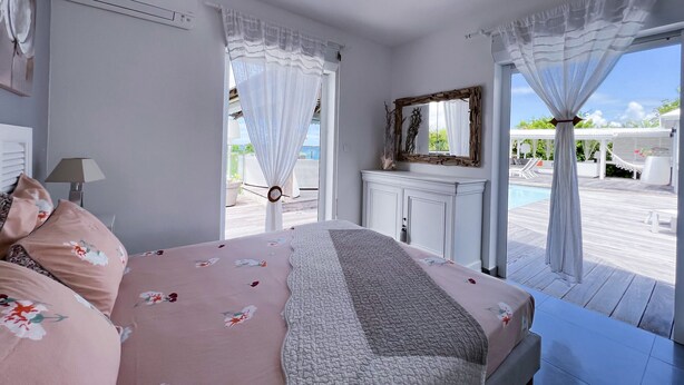 A bedroom with a bed, a bedside table, a mirror, and a view of an outdoor swimming pool. - Villa Panorama Marin · Bois Jolan · 5 BR · 426969 (Sainte-Anne)