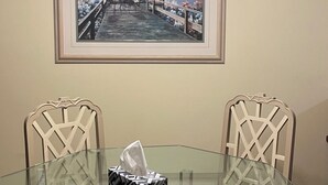 A dining area with a glass table, two chairs, a framed painting, and a chandelier. - Beautiful condo ocean view. (New Smyrna Beach)