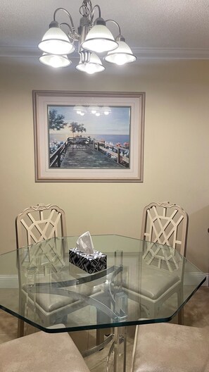 A dining area with a glass table, two chairs, a framed painting, and a chandelier. - Beautiful condo ocean view. (New Smyrna Beach)