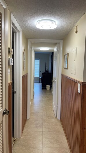 A hallway with a ceiling light, wooden paneling, a trash can, and a door leading to a room. - Beautiful condo ocean view. (New Smyrna Beach)