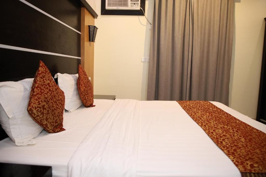 A hotel room with a bed, pillows, a headboard, a window with curtains, and a wall-mounted air conditioning unit.