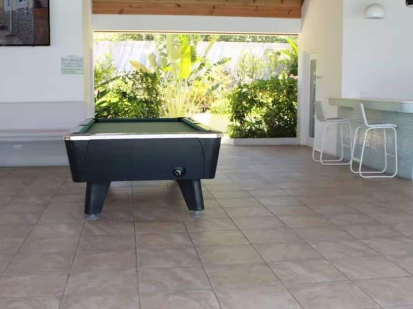 Una mesa de billar en una habitación con piso de baldosas y vistas a zonas verdes al exterior.