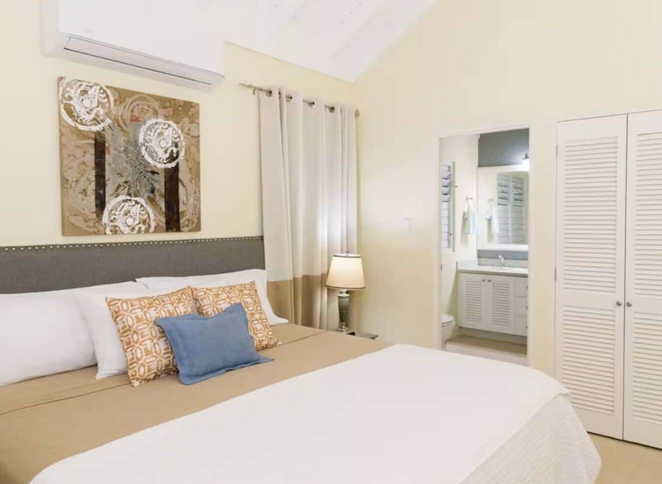 Quarto de hotel com cama, mesa de cabeceira, luminária, banheiro com espelho e guarda-roupa.