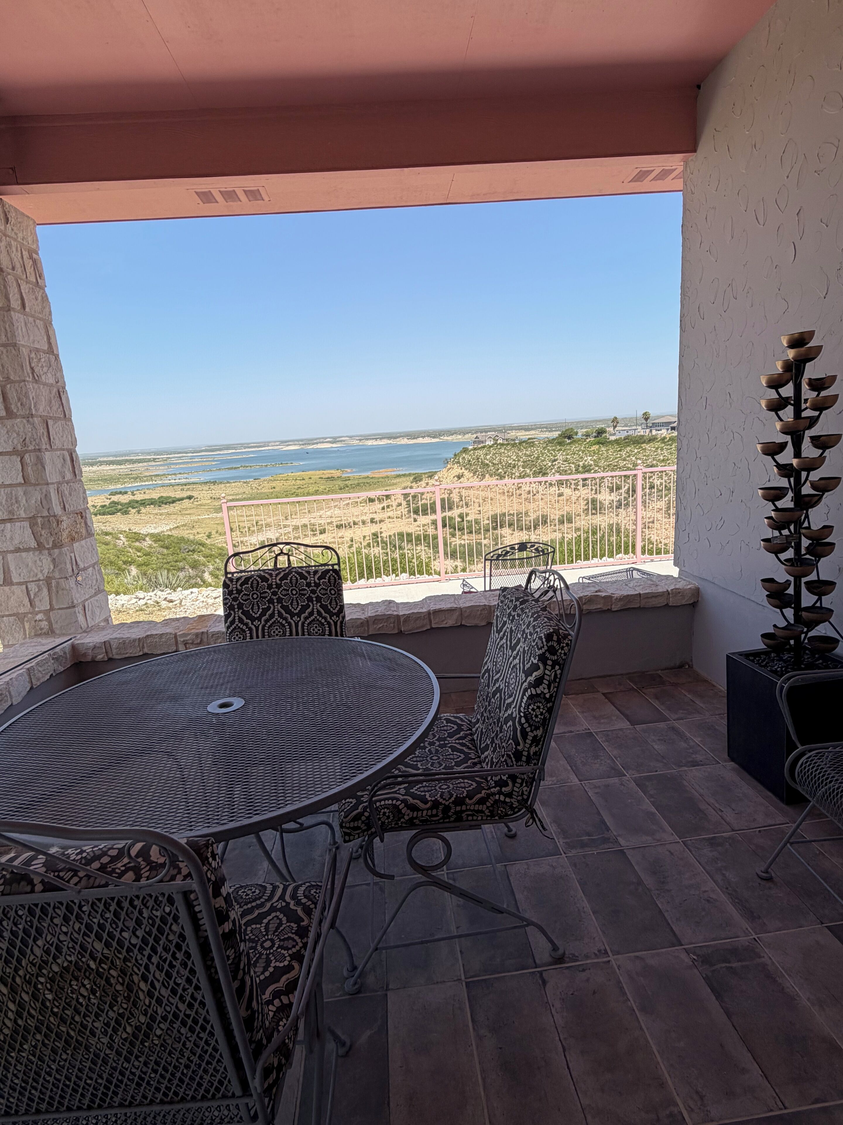 A patio with a round table and chairs, a decorative plant, and a view of a coastal landscape.